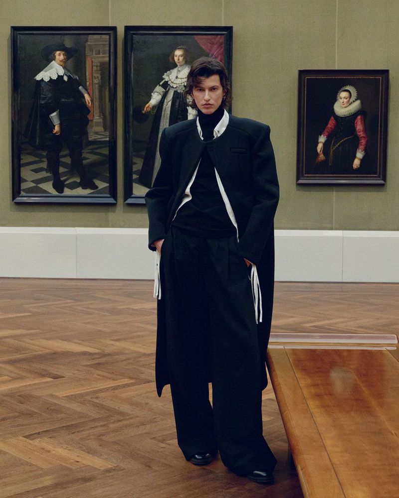 Adult male posing in a black suit and coat in front of three classic portraits displayed in a gallery with herringbone wood floors.