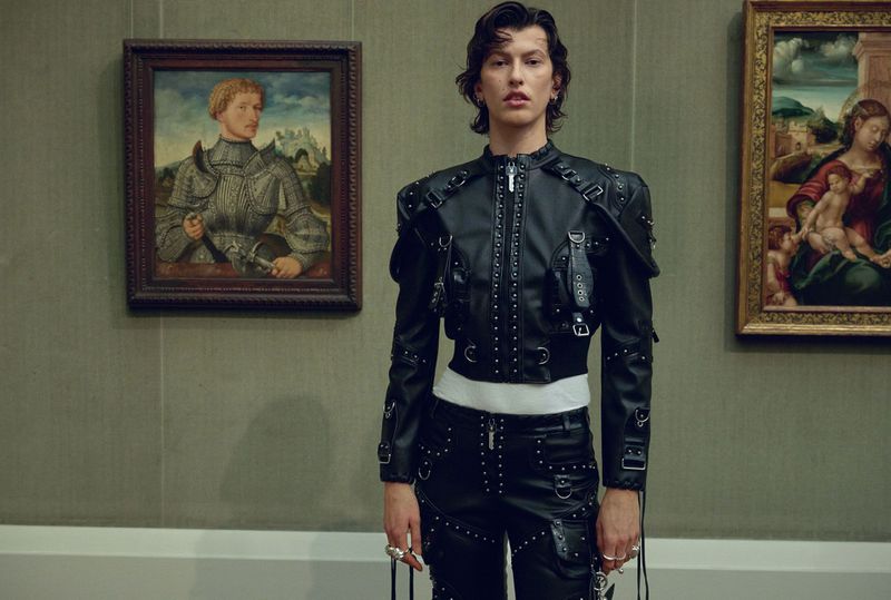 A person in black studded leather posing in front of framed paintings at a museum or art gallery; fashion editorial photography.