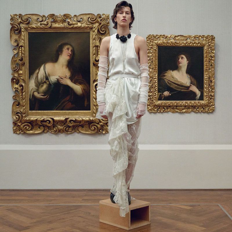 A woman in a white gown stands on a wooden box in an art gallery, two classical paintings in ornate gold frames hang behind her.