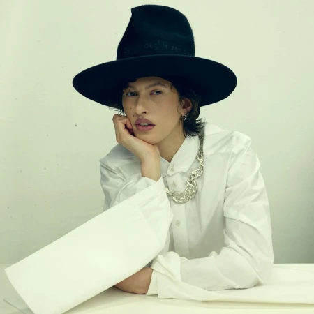 Model wearing a wide-brimmed black hat, structured white oversized shirt, and silver collar necklace resting chin in hand.