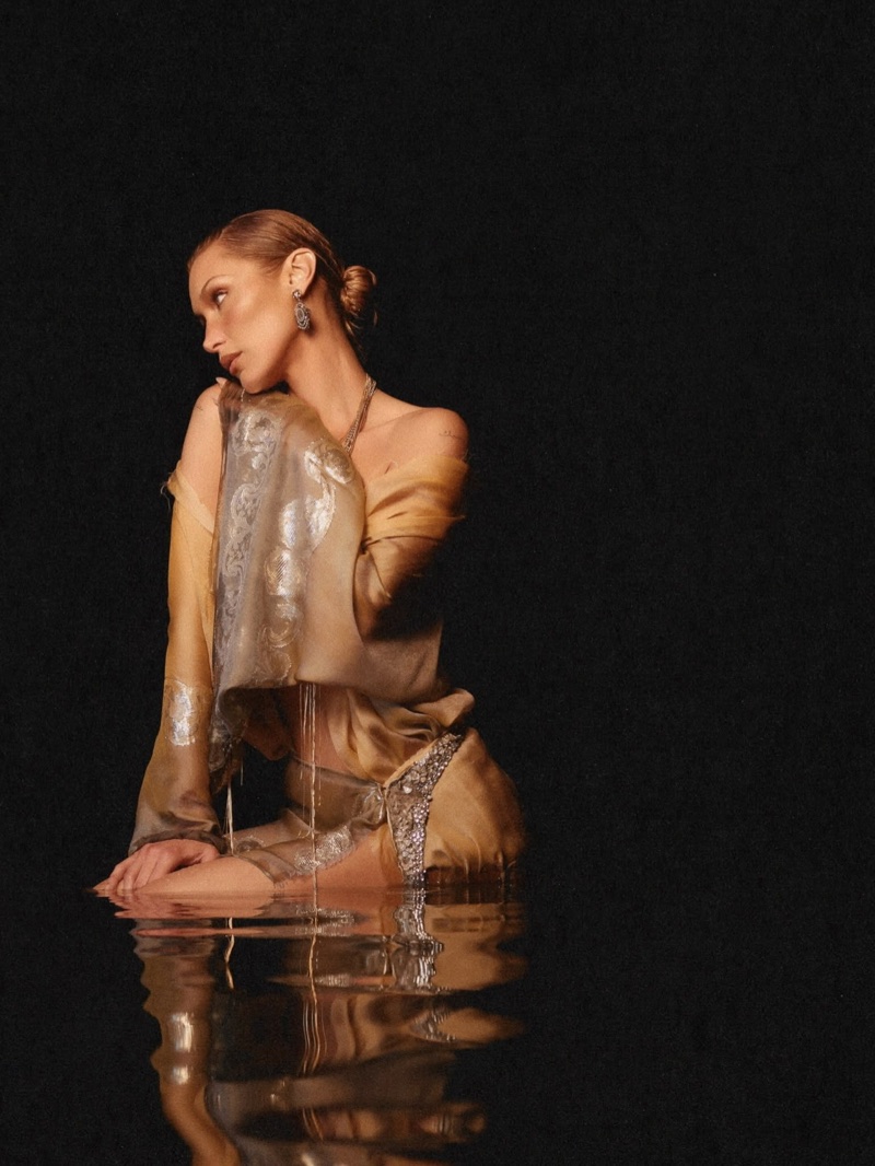 Bella Hadid posing in a silky draped garment with metallic accents, reflected in water against a dark background for Orebella