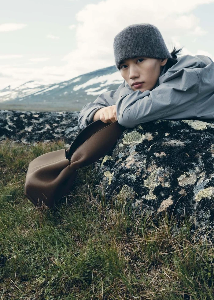 Model wearing a grey beanie and jacket leaning over a lichen-covered rock holding a brown leather bag outdoors in a mountain landscape.