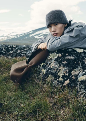 Model wearing a grey beanie and jacket leaning over a lichen-covered rock holding a brown leather bag outdoors in a mountain landscape.