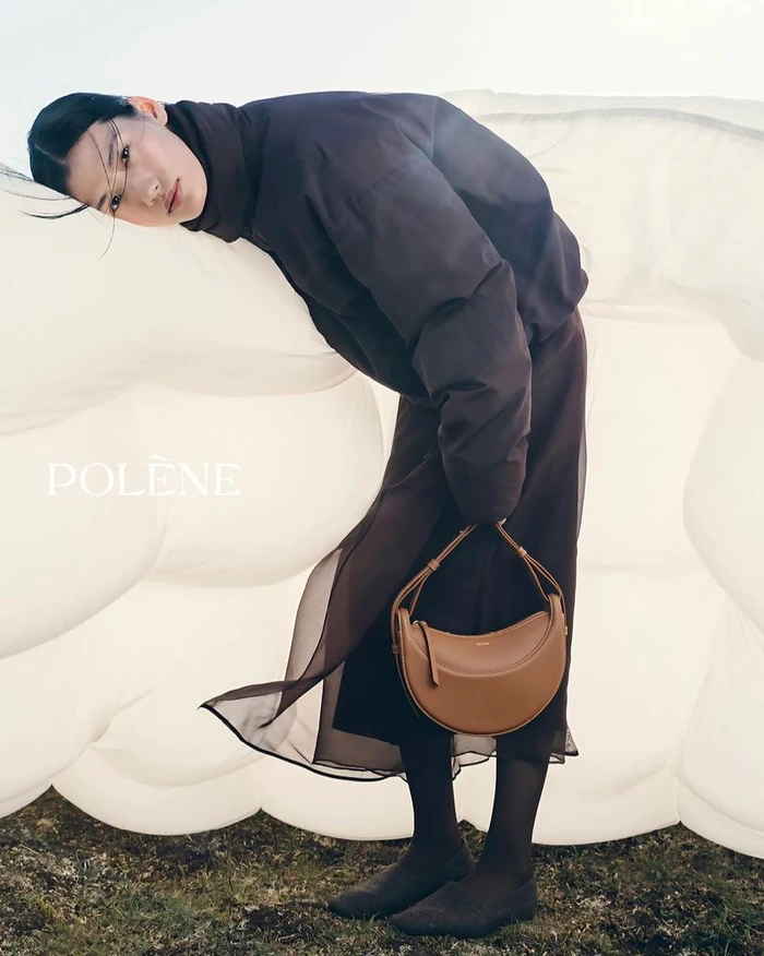 Model leaning against a large white inflatable sculpture holding a tan leather shoulder bag outdoors