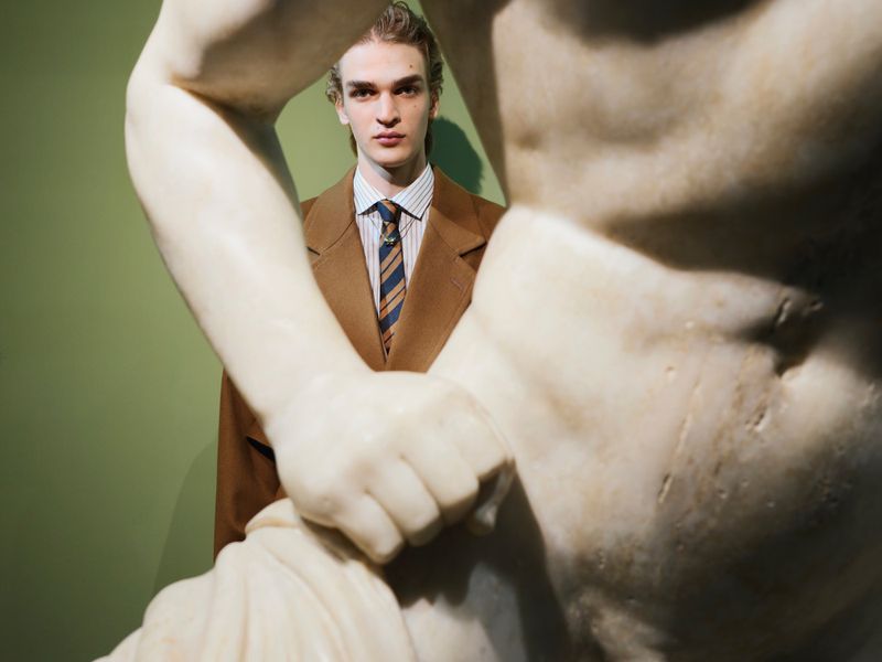 Model wearing a brown coat and striped tie framed behind a marble classical statue sculpture