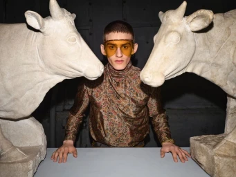 Model wearing amber-tinted visor and patterned turtleneck standing flanked by two stone cow statues.