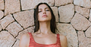 Model Irina Shayk with wet dark hair wearing a red textured top against a stacked stone wall background for Hunza G Resort 2027.