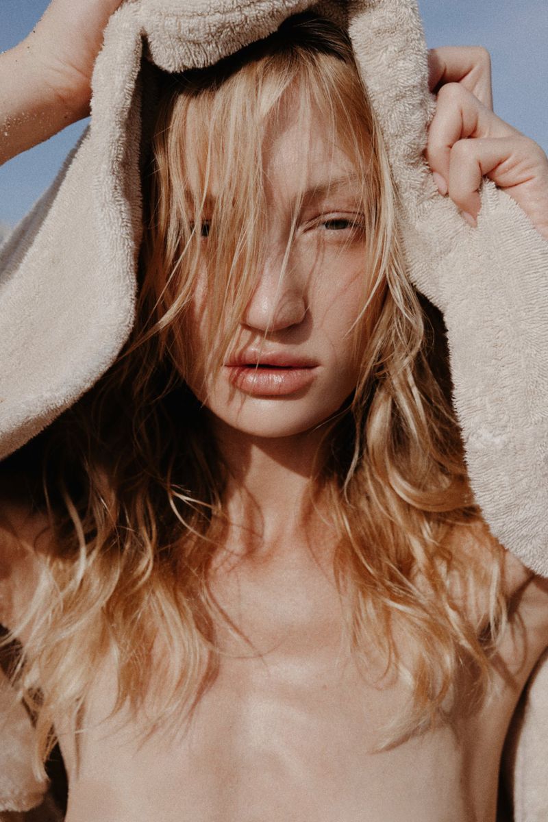 Close-up portrait of a blonde model with wet hair holding a beige towel over her head against a clear sky