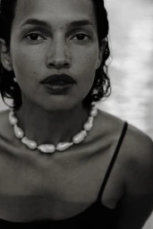 Close-up portrait of a woman wearing a chunky baroque pearl necklace and a dark strap top against a textured background.