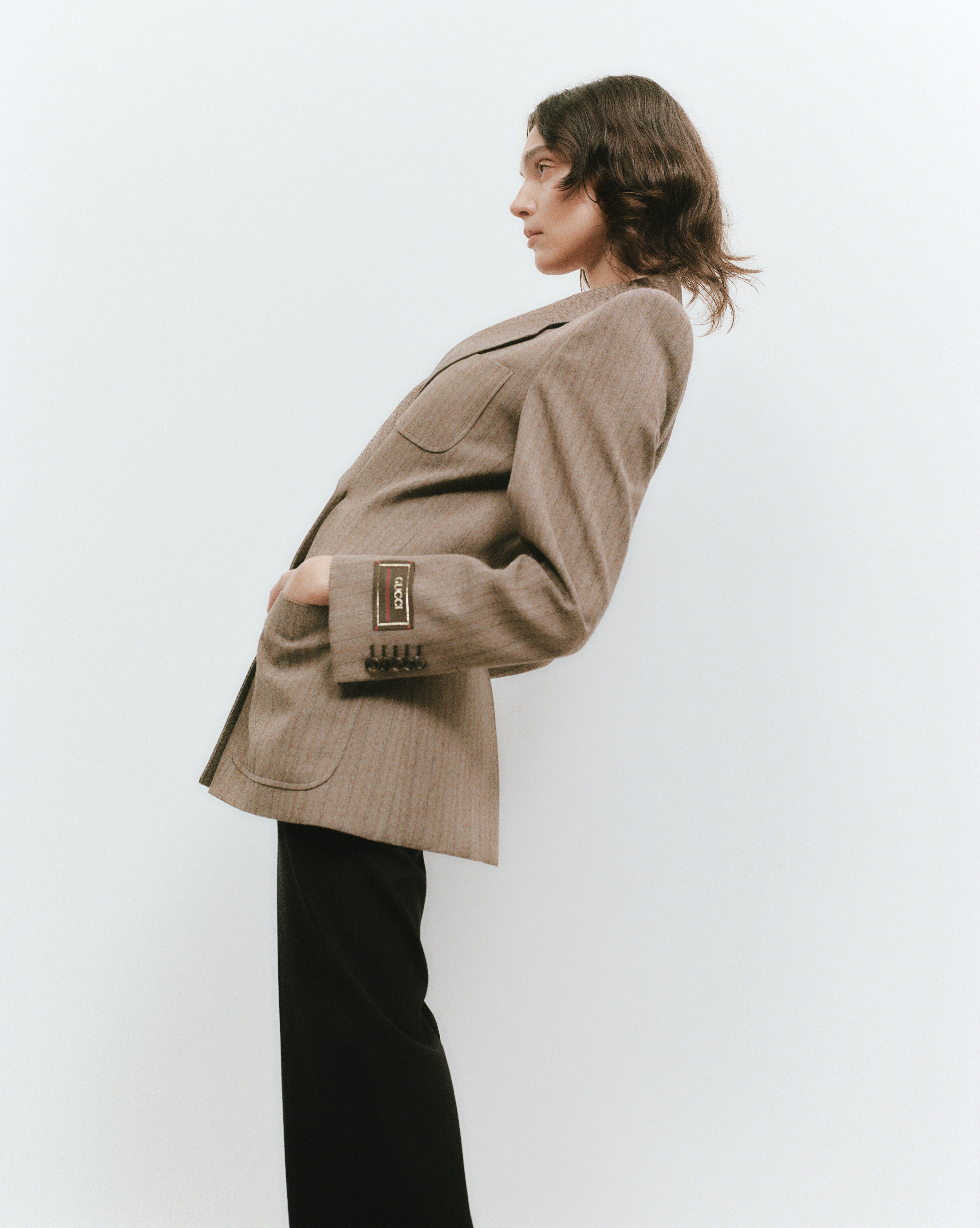 A woman in a tan herringbone blazer and black pants poses against a plain white background in a fashion editorial shot.