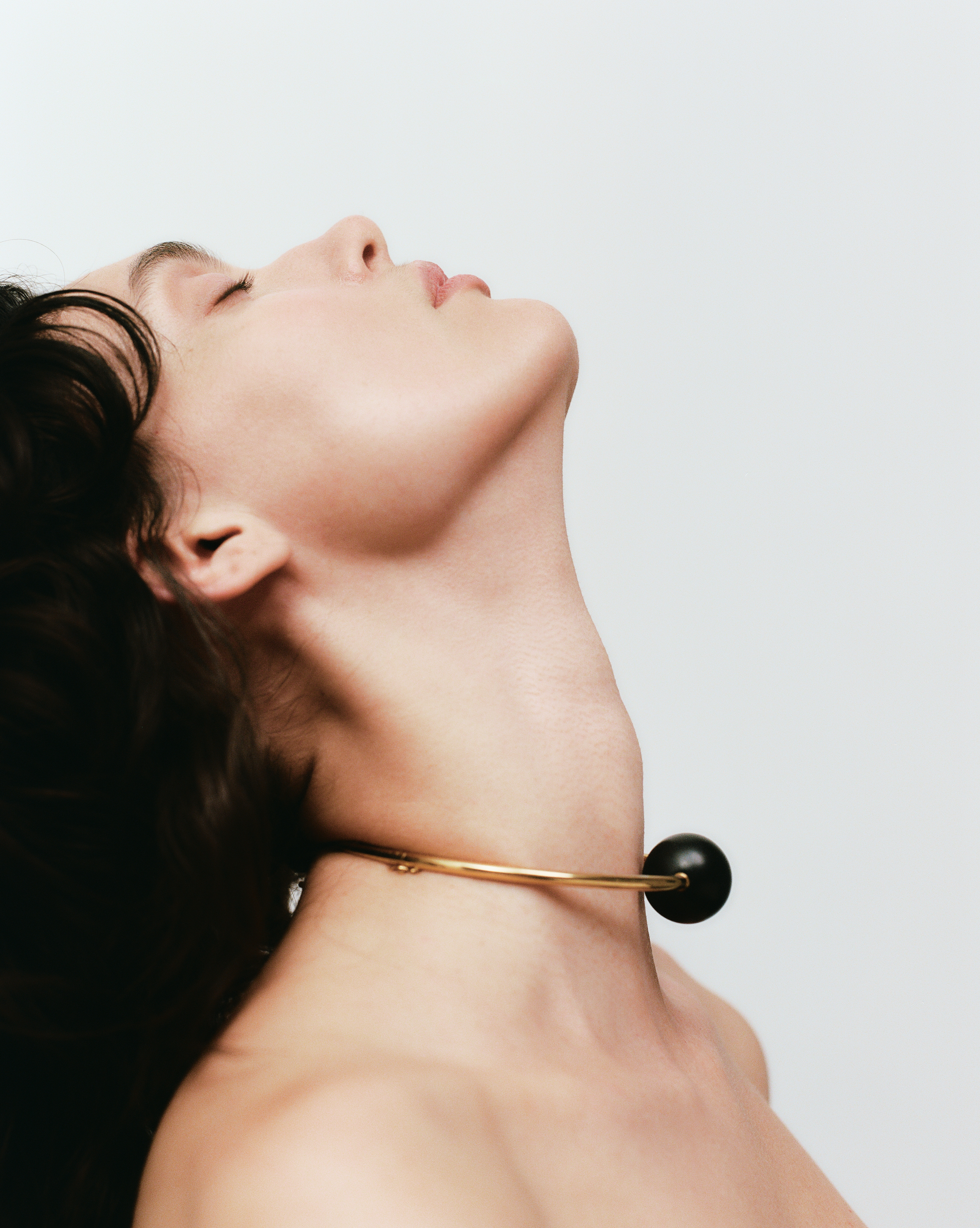 Close-up studio shot of a woman with dark hair and a golden choker with a black sphere, against a clean white background.
