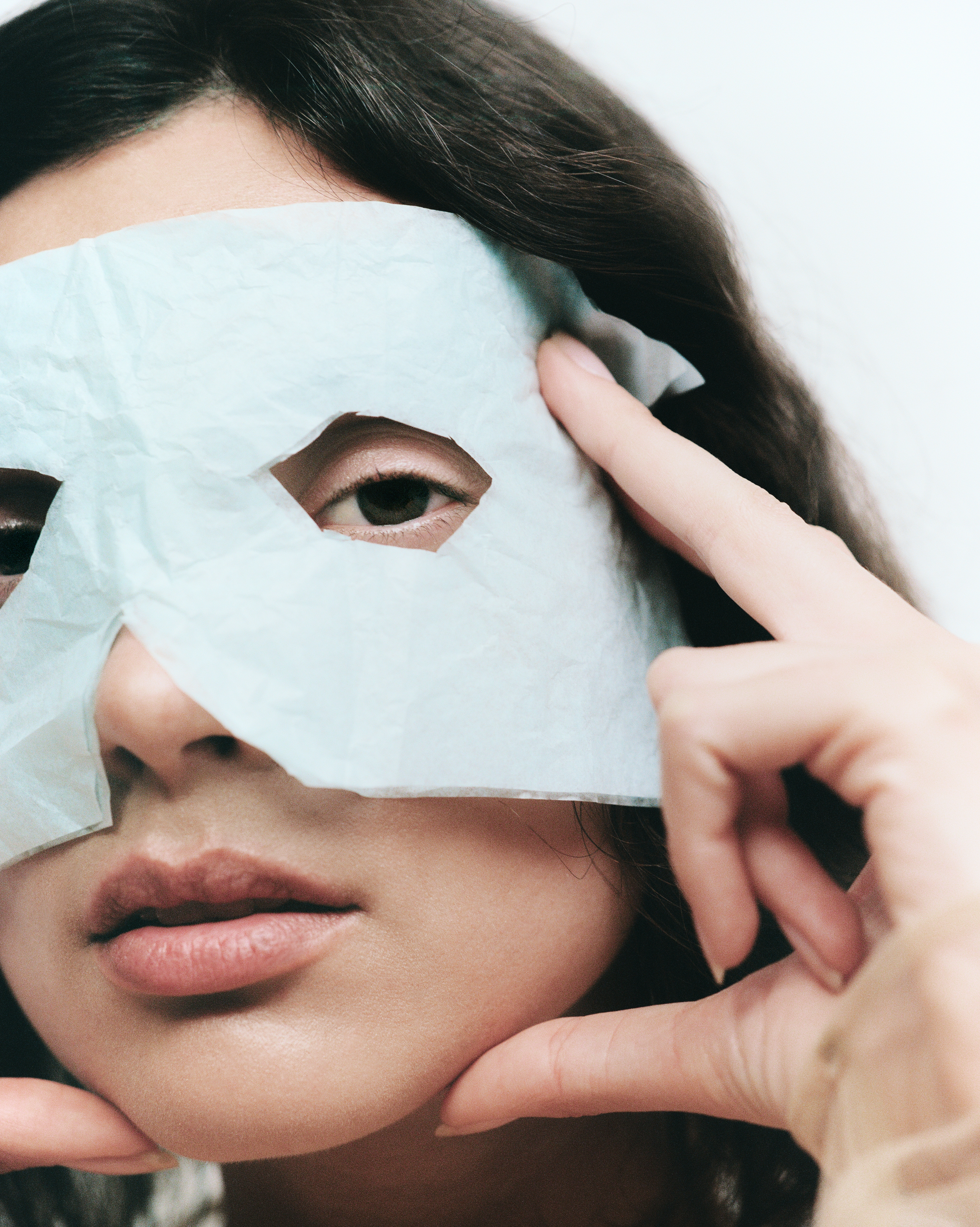 Close-up shot of a woman with a blue facial mask on, holding her fingers near her face, against a white background in a studio.