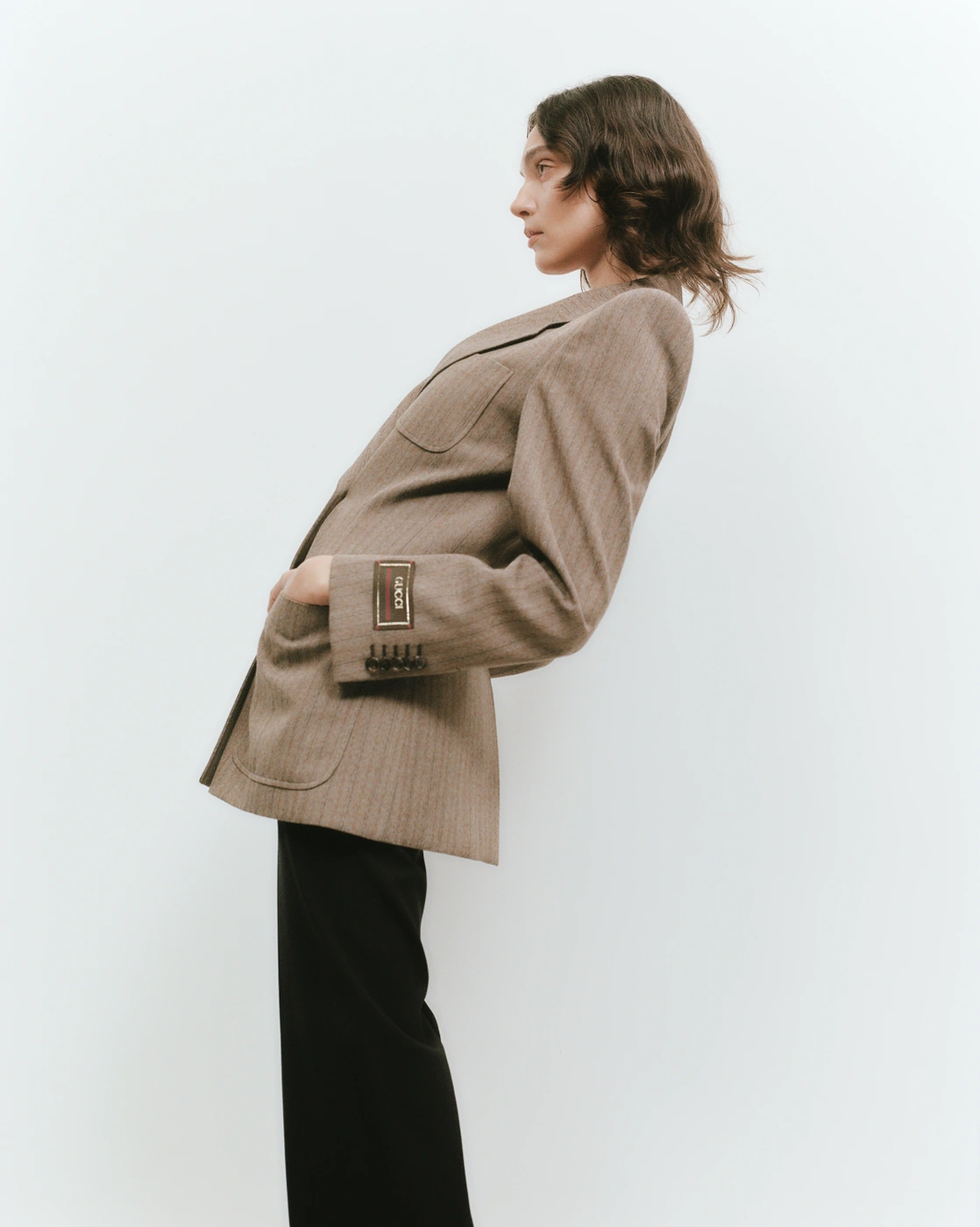 Model wearing a brown blazer and black trousers poses against a plain white background for ONE Magazine.