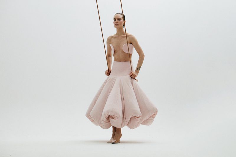 A slender woman in a pink gown strikes a pose, hands clasped on a brown rope swing, set against a clean, stark white backdrop.