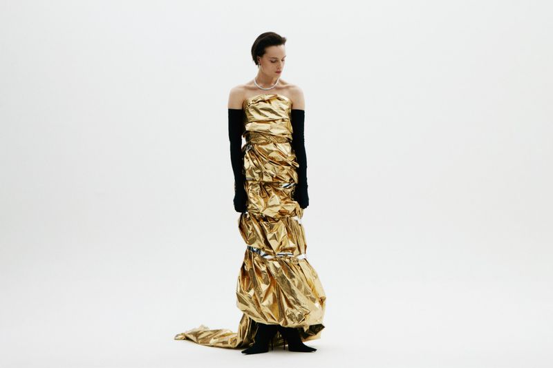 Woman in a gold dress and black gloves stands in a white photo studio. The dress is metallic and shiny, catching the light.