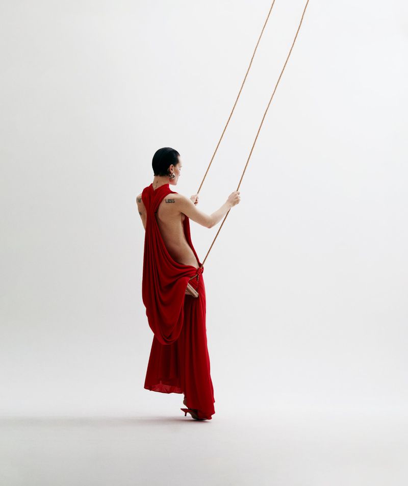 Woman in a red draped gown standing on a wooden swing with rope, posing in a brightly lit studio setting with a white background.