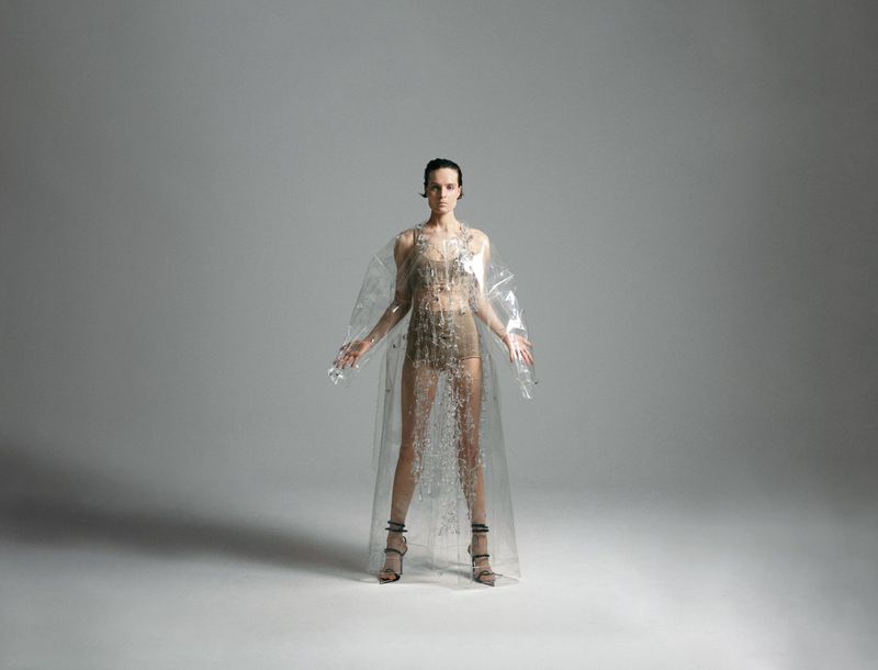 Full shot of a young woman in a sheer plastic coat and heels posing against a neutral gray background in a studio setting. 