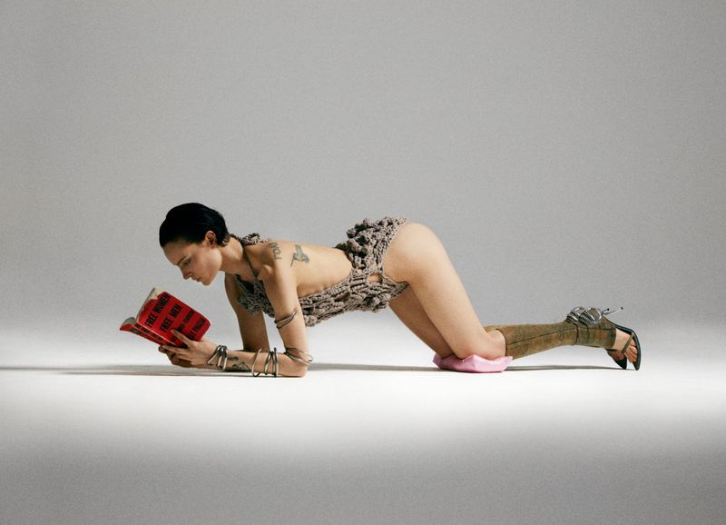 An adult woman poses in a crocheted bodysuit while reading a book on a white background. She is accessorized with stacked silver bangles.