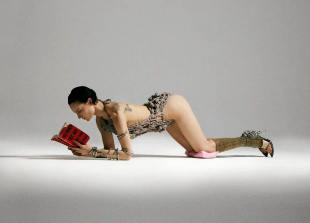 An adult woman poses in a crocheted bodysuit while reading a book on a white background. She is accessorized with stacked silver bangles.