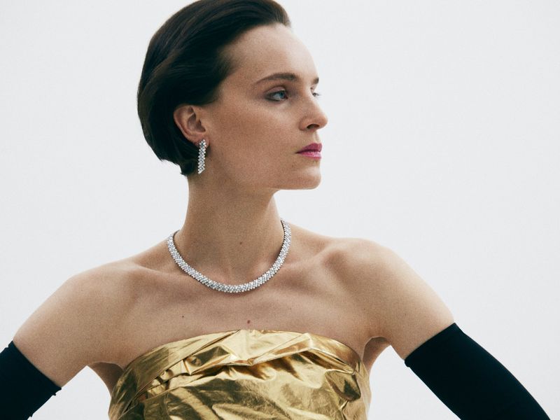 Elegant woman in gold strapless top wearing diamond necklace and earrings against a clean white backdrop in a fashion shot.