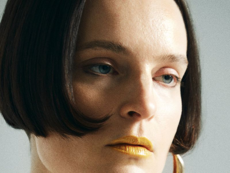 Close-up portrait of an adult female with short, dark hair and gold lipstick, set against a plain grey background, with subtle jewelry.