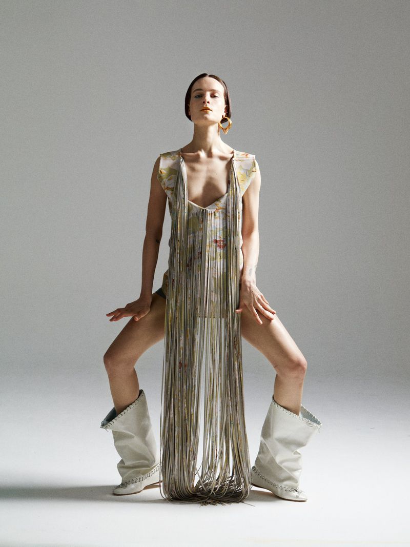 Fashion shot of a woman in a patterned dress with long fringe, gold hoop earrings, and white heeled boots posing against a light gray background.