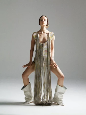 Fashion shot of a woman in a patterned dress with long fringe, gold hoop earrings, and white heeled boots posing against a light gray background.