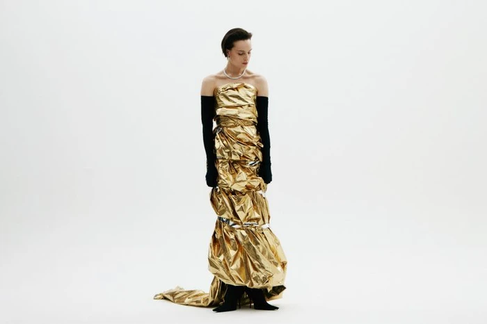 Model in metallic gold foil strapless gown wearing long black opera gloves against a seamless white background.