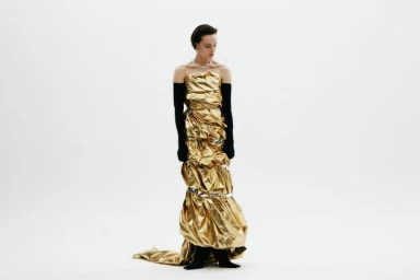Model in metallic gold foil strapless gown wearing long black opera gloves against a seamless white background.
