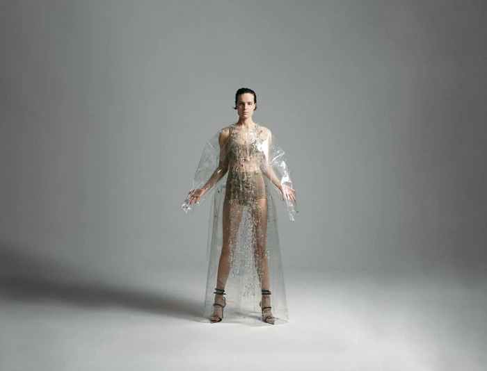 Model standing centrally in a clear plastic cape and strappy heels against a seamless grey backdrop.