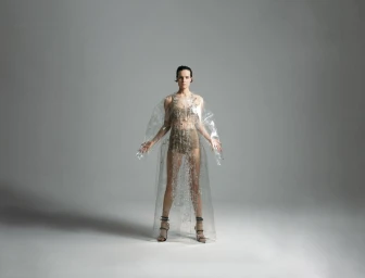 Model standing centrally in a clear plastic cape and strappy heels against a seamless grey backdrop.