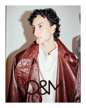 Model with dark curly hair wearing a deep red leather coat over a white top against a plain wall.