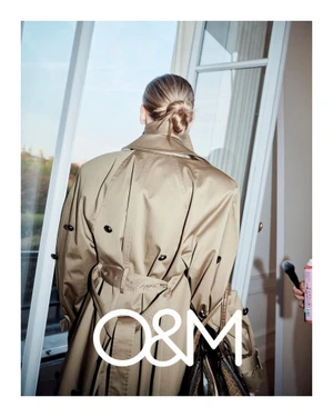 Model seen from behind wearing a tan trench coat standing near a bright window for O and M Hair photography by Bridget Fleming