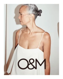 Older woman with grey hair in a bun wearing a white slip top against a plain studio background for O&M Hair