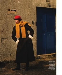 Model wearing a red hat, yellow scarf, and long dark coat standing outdoors near a blue door.
