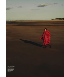 Model standing on a vast sandy beach wearing a long red plaid coat under a wide horizon and overcast sky.