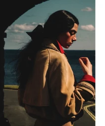 Profile of a woman with dark hair in a tan coat with red collar standing outdoors near the ocean.