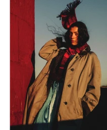 Model wearing a tan coat, red scarf, and patterned headpiece outdoors near a red structure.