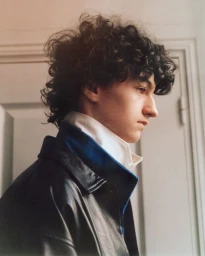 Profile portrait of a young man with dark curly hair wearing a dark leather jacket and white collar, indoors.