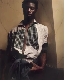 Black man wearing a patchwork plaid shirt leaning against an architectural wall in dim lighting