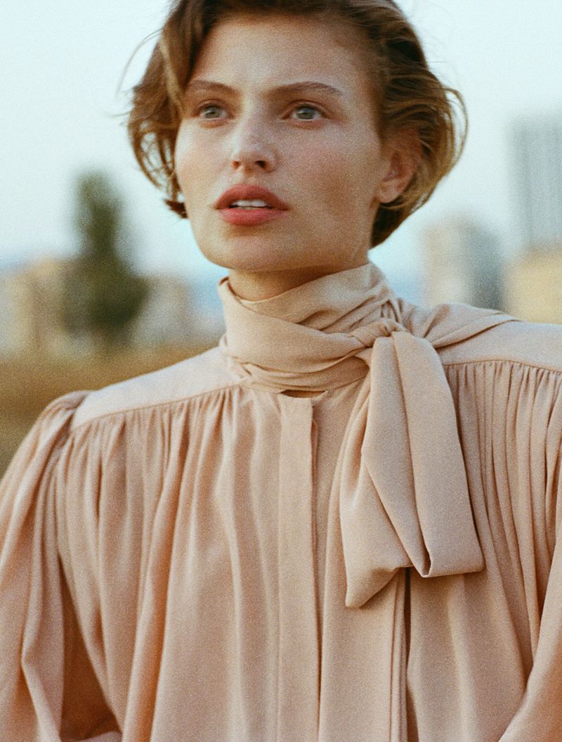 Portrait of an adult female with short brown hair wearing a peach-colored tie-neck blouse, against a blurred city background.