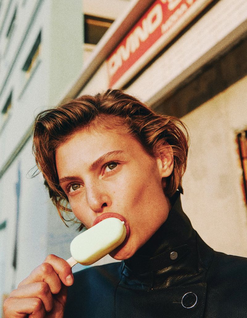 Adult female model in outdoor fashion shot eating a lemon ice cream bar in front of a building with awning, medium portrait.