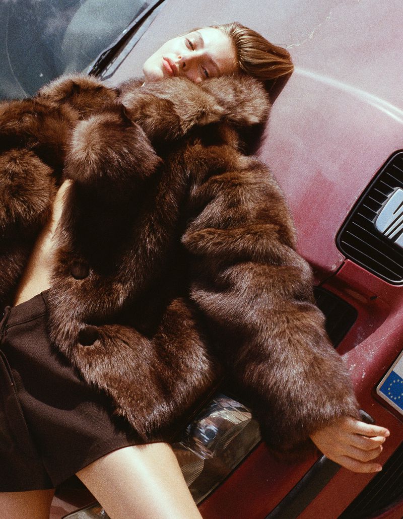 A woman in a brown fur coat reclines on a red vintage car. Shot outdoors in daylight with a shallow depth of field. Fashion editorial.