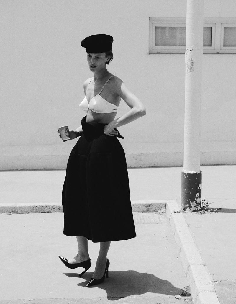 Model Bibi Breslin wearing a black beret, white bra top, black voluminous midi skirt, and pointed heels outdoors beside a white pole.