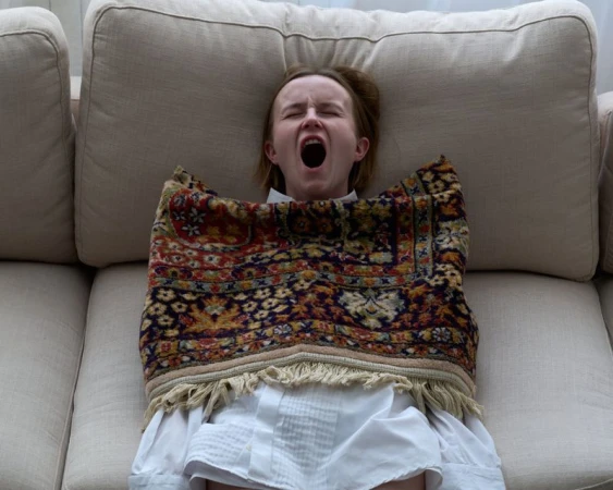 Woman wearing a white shirt and patterned fringed rug lying back on a light beige sofa with her mouth open