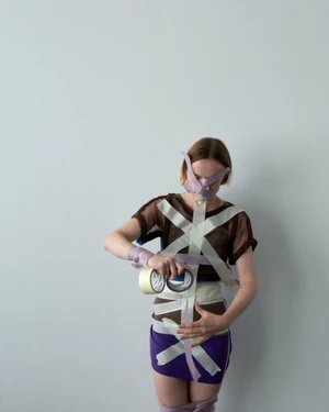 Woman in brown mesh top and purple skirt applying adhesive tape to her body in a studio setting
