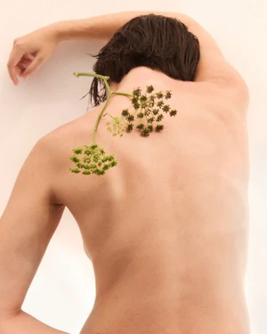 Close-up of a person's bare back with green flowering plant stems resting across the shoulders against a plain light background