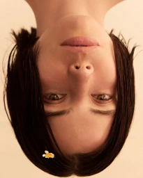 Inverted close-up portrait of a woman with dark bobbed hair and a small white flower tucked behind her ear against a neutral background.