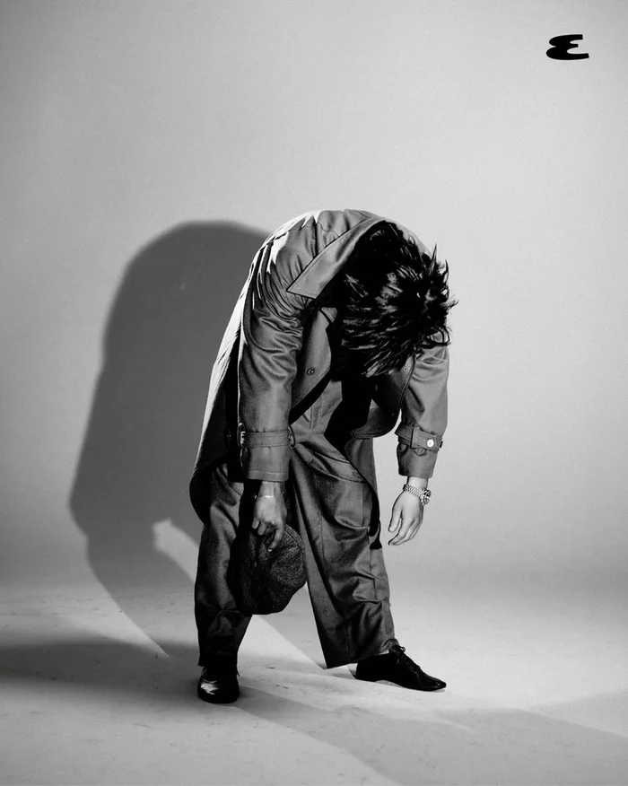Glen Powell bent over wearing a trench coat and holding a cap against a light grey seamless studio background.