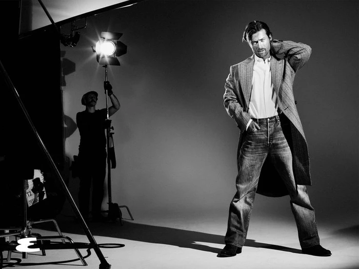Glen Powell in a patterned coat and jeans posing in a studio with a lighting crew visible in the background.